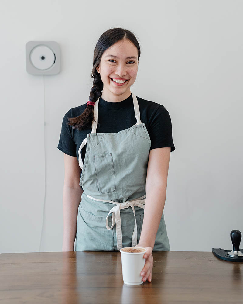 happy ethnic female barista offering coffee in paper cup | apron pattern